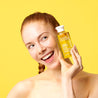 Woman holding the Vitamin C Cleansing Oil next to her face and smiling while looking away.