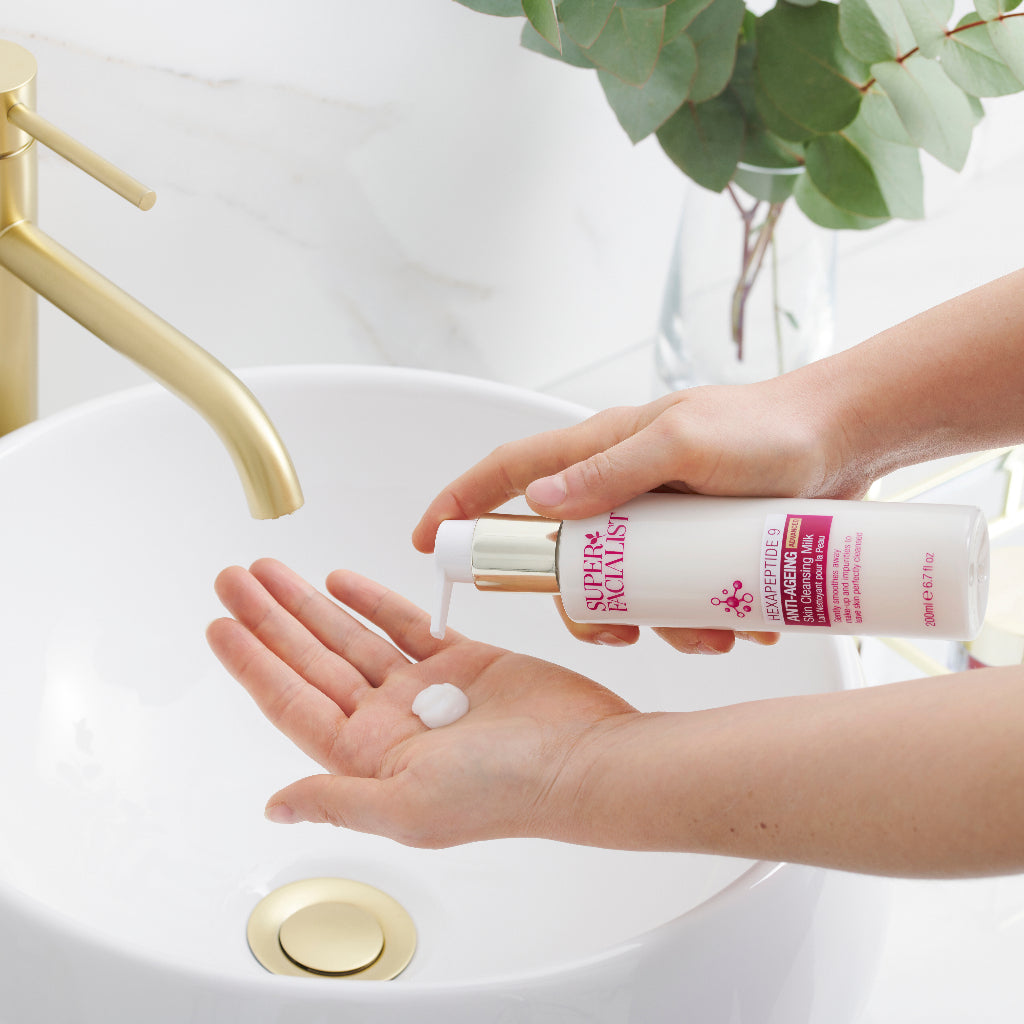 Hand pumping out cleansing milk onto palm of another hand above a white sink with a gold tap