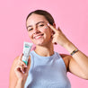 Teen model applying Moisturiser onto her cheek with one finger against pink background