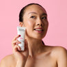 Model holding white day cream tube next to her face while looking away against a pink background