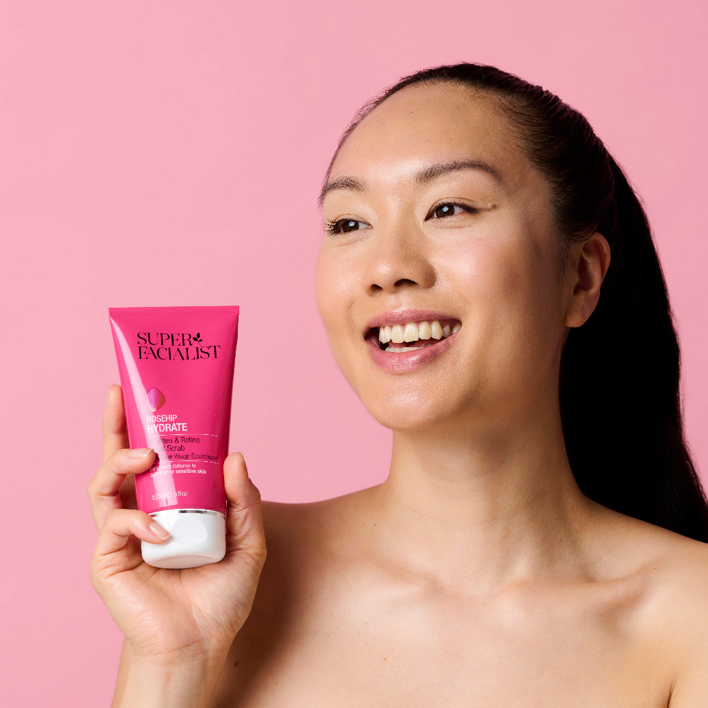 Smiling model holding the rosehip facial scrub in front of a pink backdrop