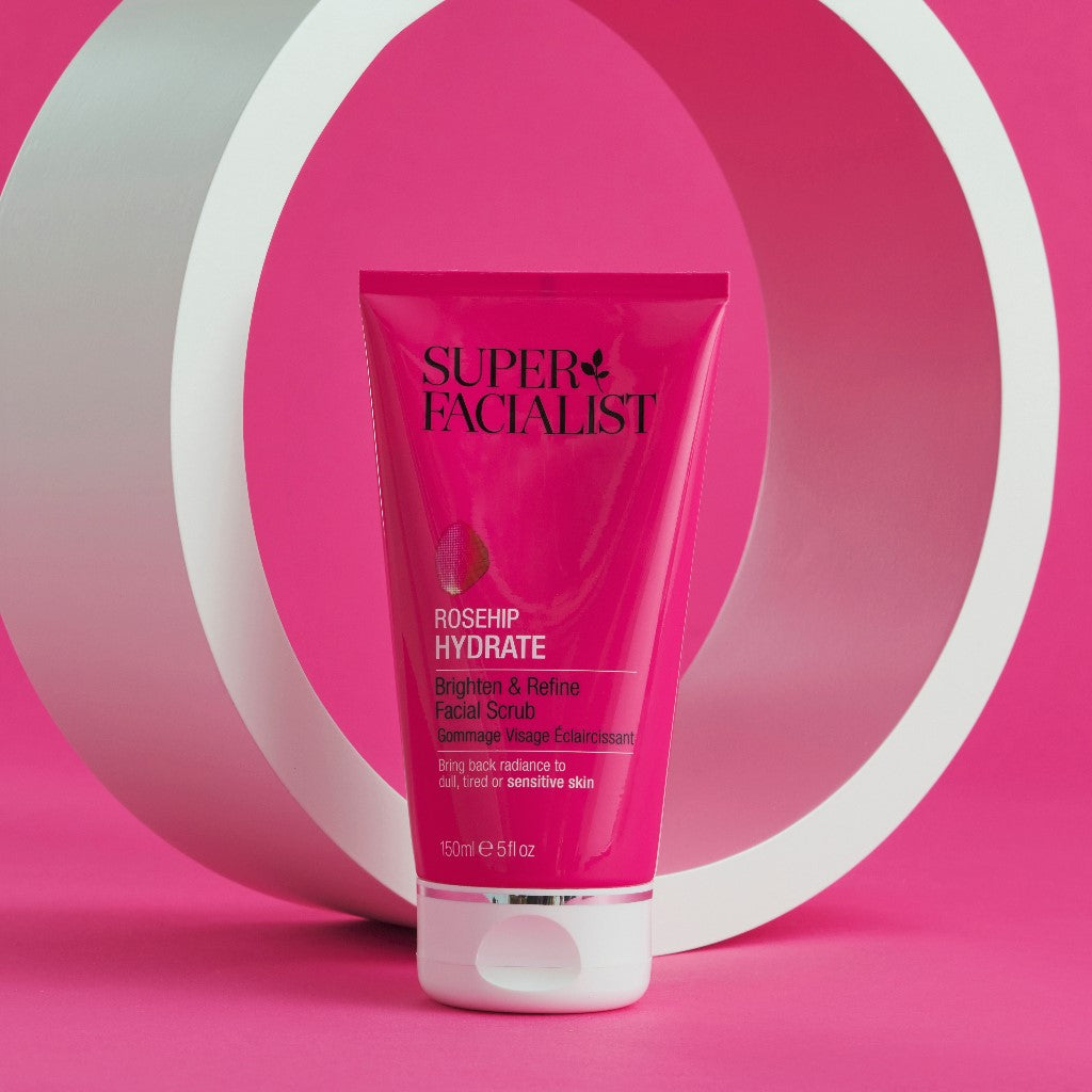 Rosehip facial scrub tube in front of a white circular prop against a dark pink backdrop