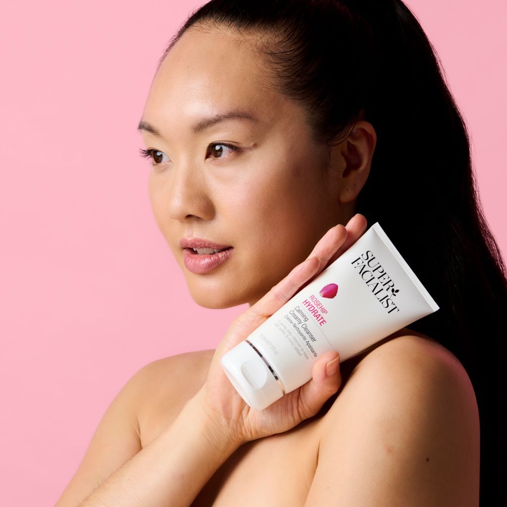 Model holding the creamy cleanser tube next to her neck against a pink background