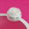 Night cream jar placed on top of a circular white prop against a dark pink background