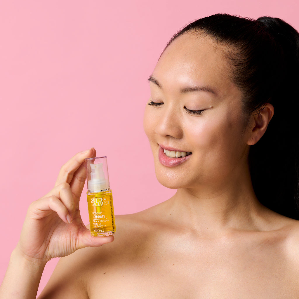 Model holding a facial oil bottle in her hand while looking at it and smiling in front of a pink backdrop