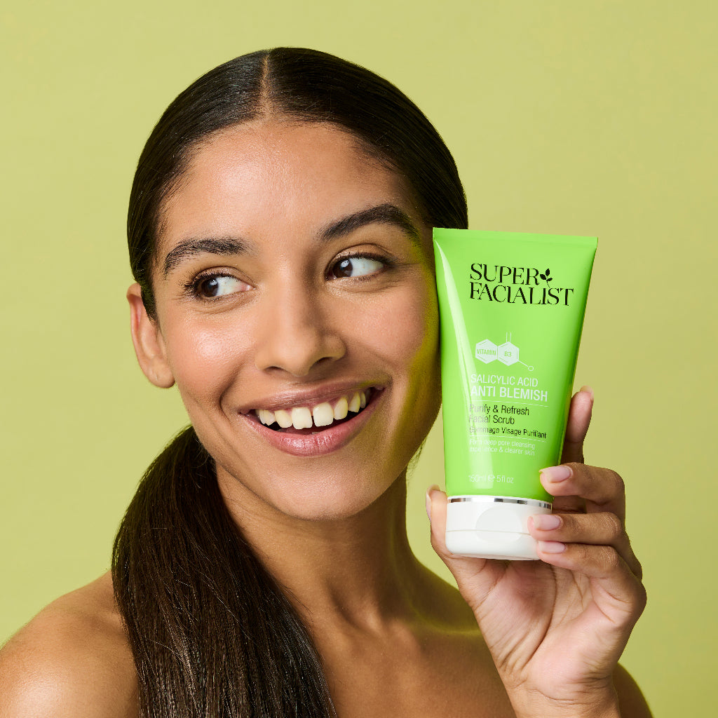 Model holding salicylic acid facial scrub green tube next to her cheek while looking the other way