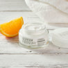 Open night cream jar next to an orange quarter on top of a white wooden table