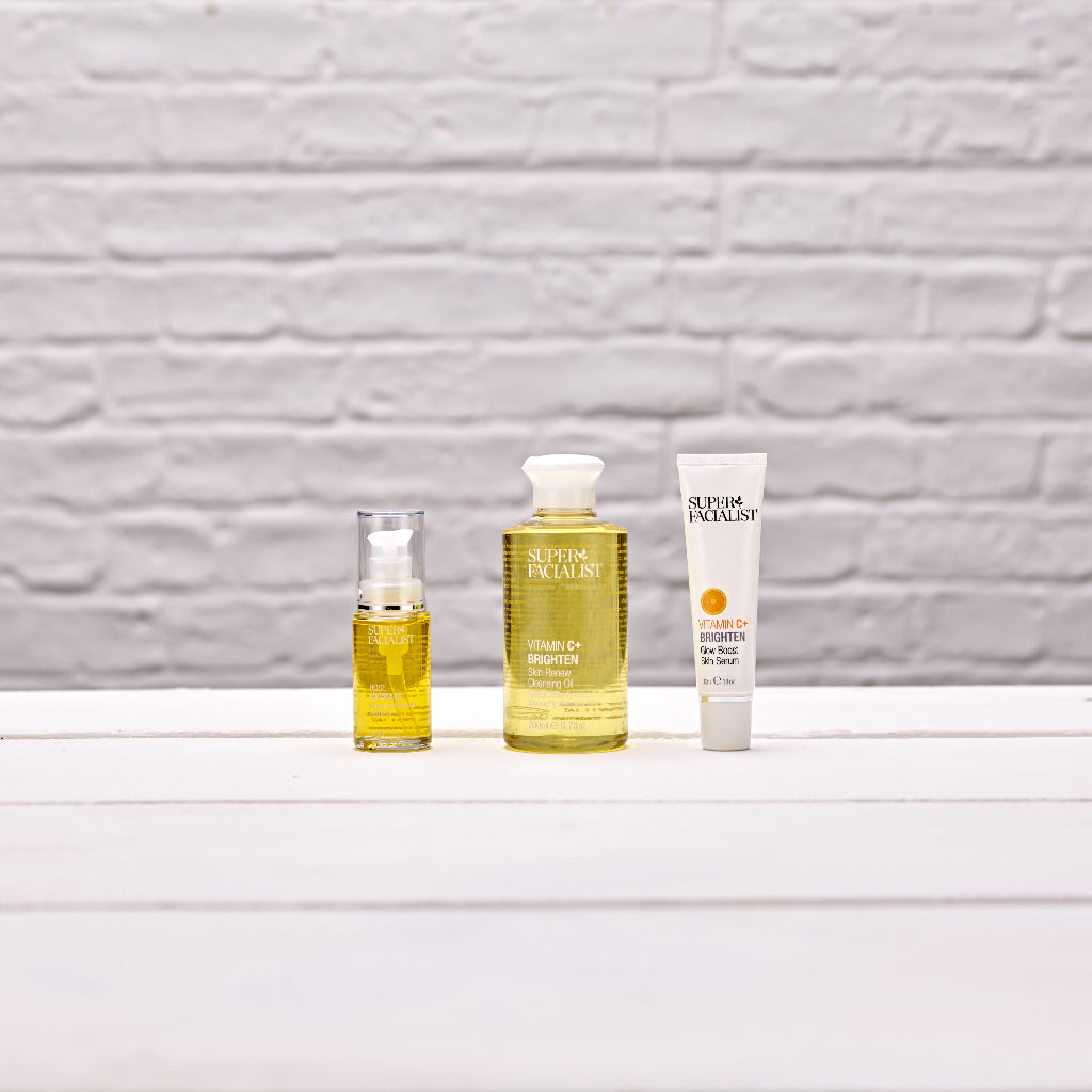 Yellow tube of rosehip facial oil next to yellow bottle of vitamin c cleansing oil next to vitamin c serum tube against a white brick wall and white wooden table